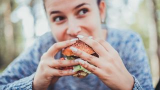 A woman is shown smiling and eating a burger looking away