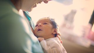 A mother is shown smiling at her newborn baby that she is holding at her breast. The baby is looking up at his face. The background is blurry.