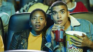 Two people are watching a movie in a theater, while one is holding a bucket of popcorn and soda