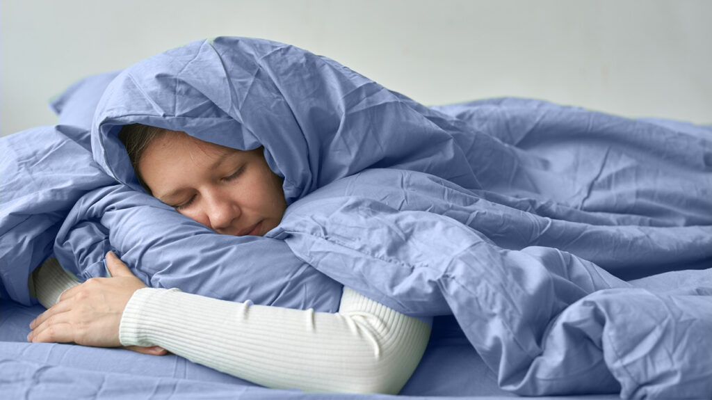 A person lying in bed wrapped in a duvet and hugging a pillow with his eyes closed