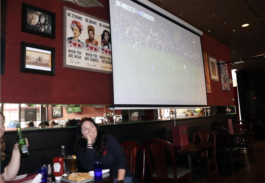 A woman sitting at a table watching a football game on a big screen in a restaurant.