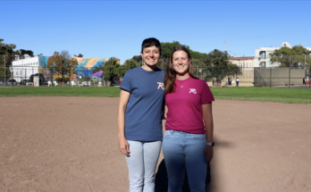 Two people are standing on a baseball field, both wearing branded t-shirts. One wears blue, the other wears red. Buildings and trees are in the background.