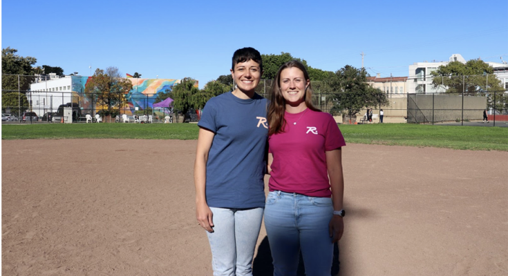 Two people are standing on a baseball field, both wearing branded t-shirts. One wears blue, the other wears red. Buildings and trees are in the background.