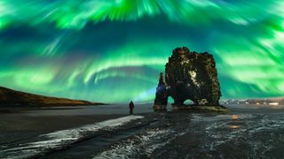 A person stands on a dark beach under a bright display of green and blue northern lights, which flash across the sky in a powerful, swirling pattern. A large, jagged rock rises nearby, adding to this amazing scene.